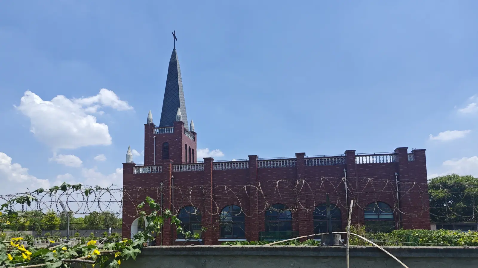 A long red-brick church with a tower and narrow pointed spire, arched windows and razor wire in the foreground, under a bright blue sky.