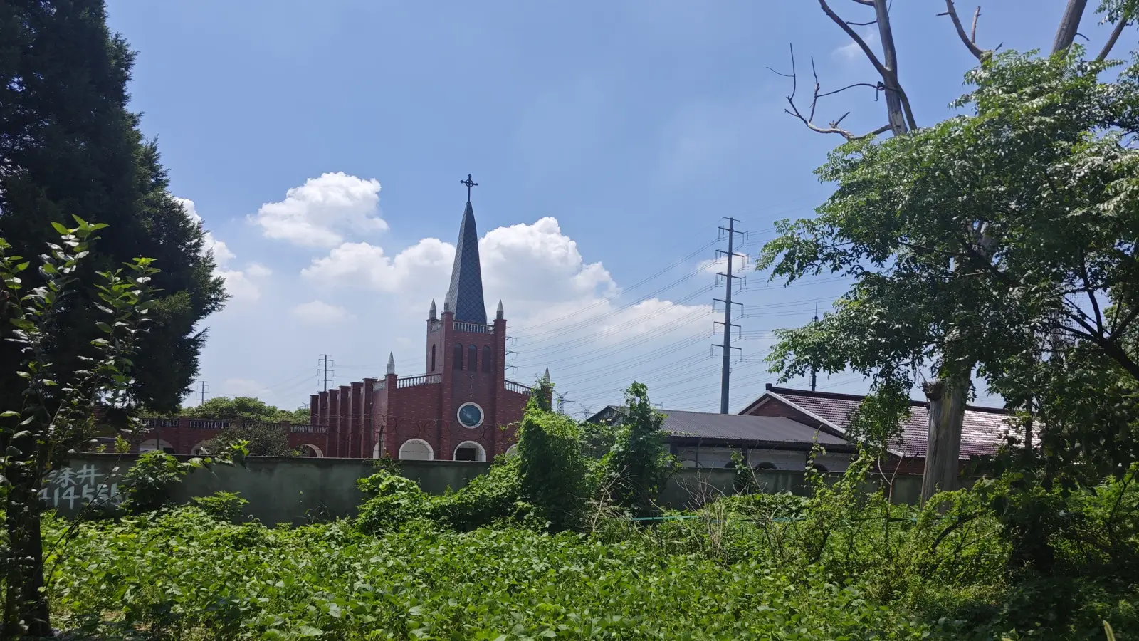 A red-brick tower with a slender dark spire beyond a green field and trees.