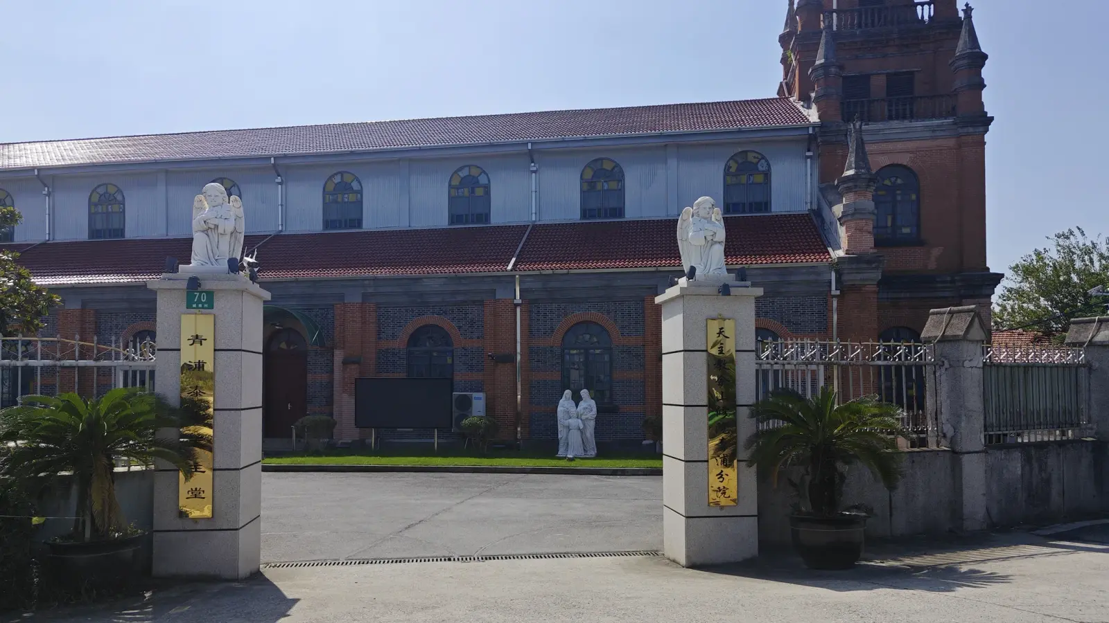 Two white gate posts with angel statues in front of a long brick church wall and tower.