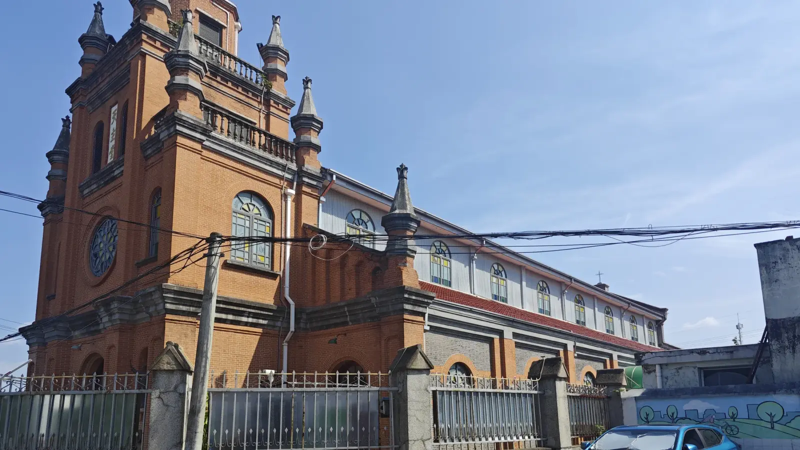 A tall orange-brick church tower with round window and pinnacles, and the nave roofline behind an iron fence.