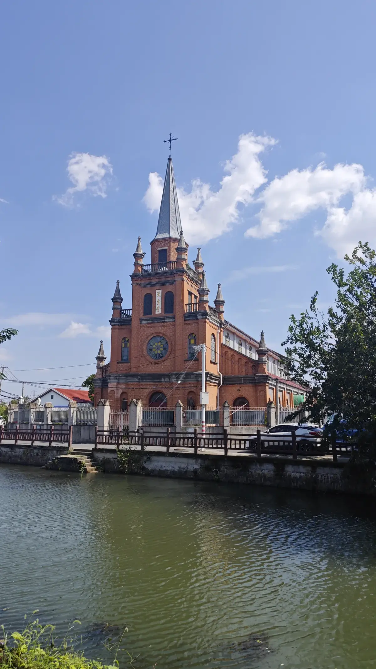 An orange-brick church with a tall central spire and corner turrets on the far side of a still pond under a blue sky.