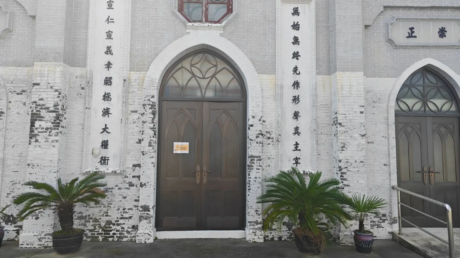 A pair of arched brown doors with Gothic tracery above, set in a grey wall with painted vertical Chinese couplets.