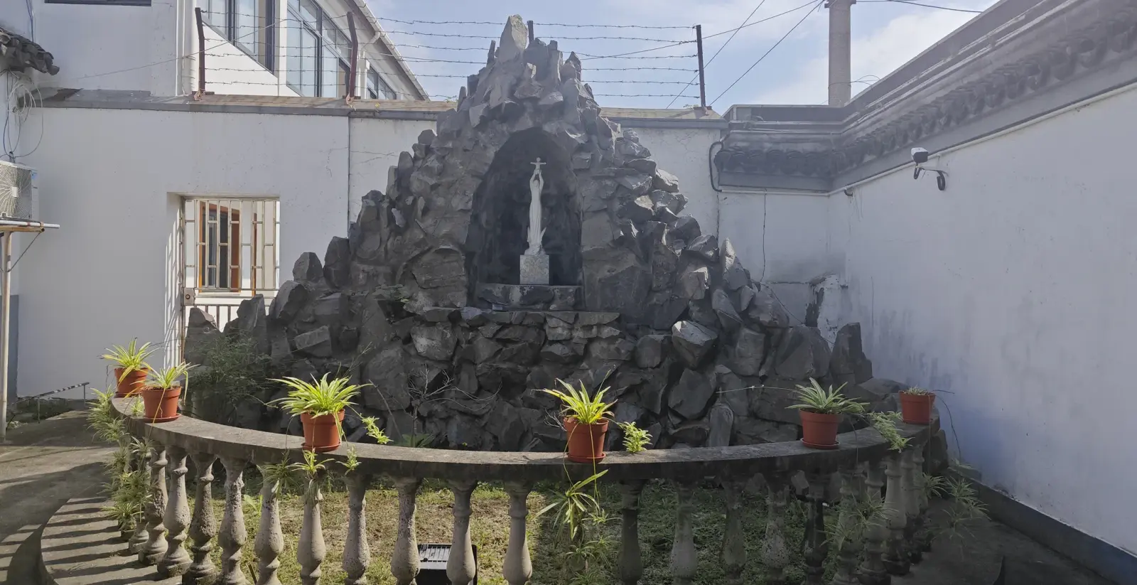 A dark stone grotto with a small white statue, surrounded by a low stone balustrade and potted red flowers.