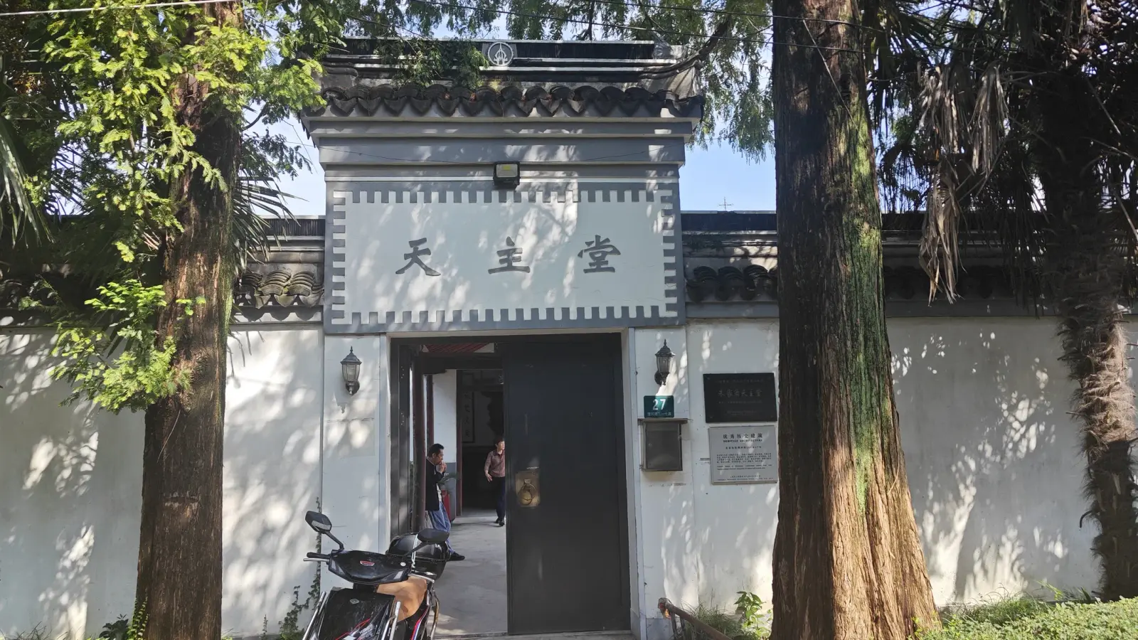 A white-walled gateway with a grey tiled roof and a sign reading 'tian zhu tang', framed by tall trees.