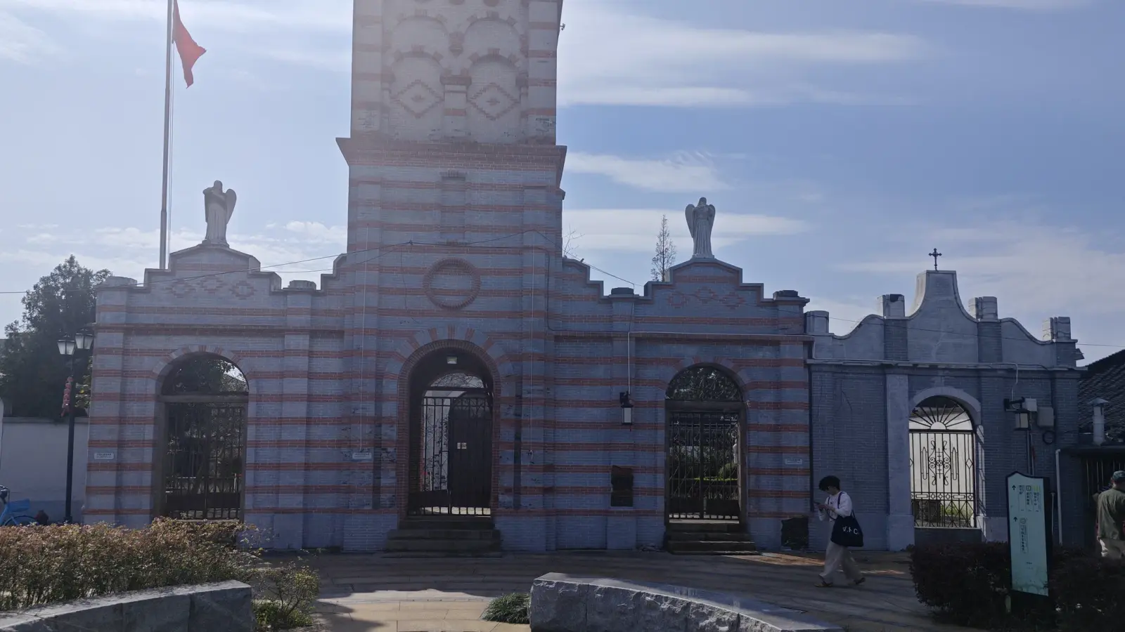 A church front of striped grey and pink masonry with a square tower, three arched openings and a flag, seen across a small plaza.
