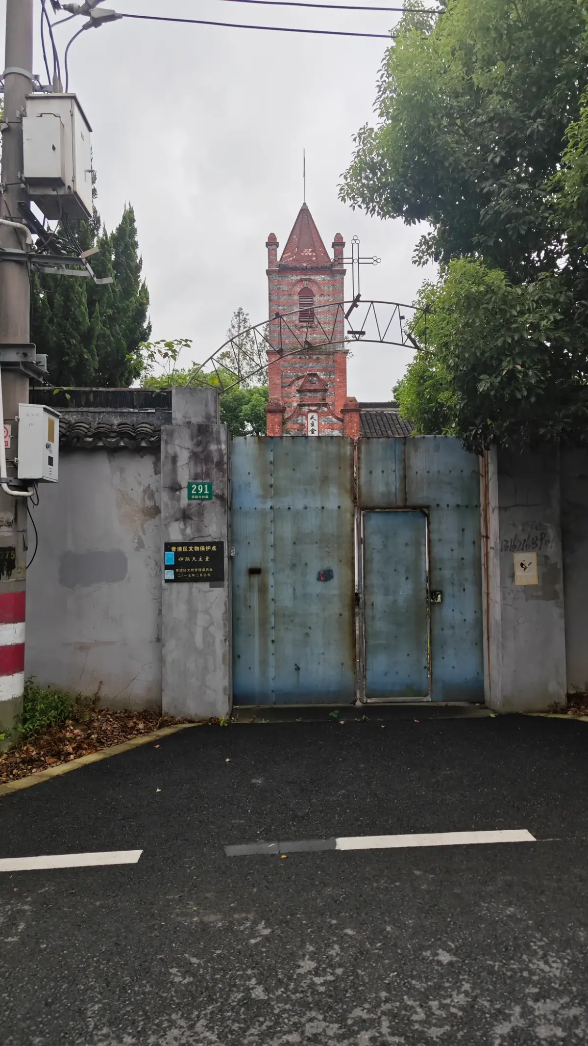 A red brick tower with a pointed top behind a blue steel gate and grey wall.