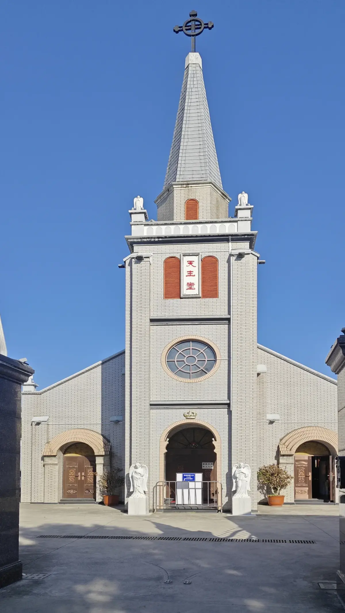 A pale church front with a tall narrow tower and spire, a round window, red shutters and small pinnacles on dark gate piers, under a blue sky.