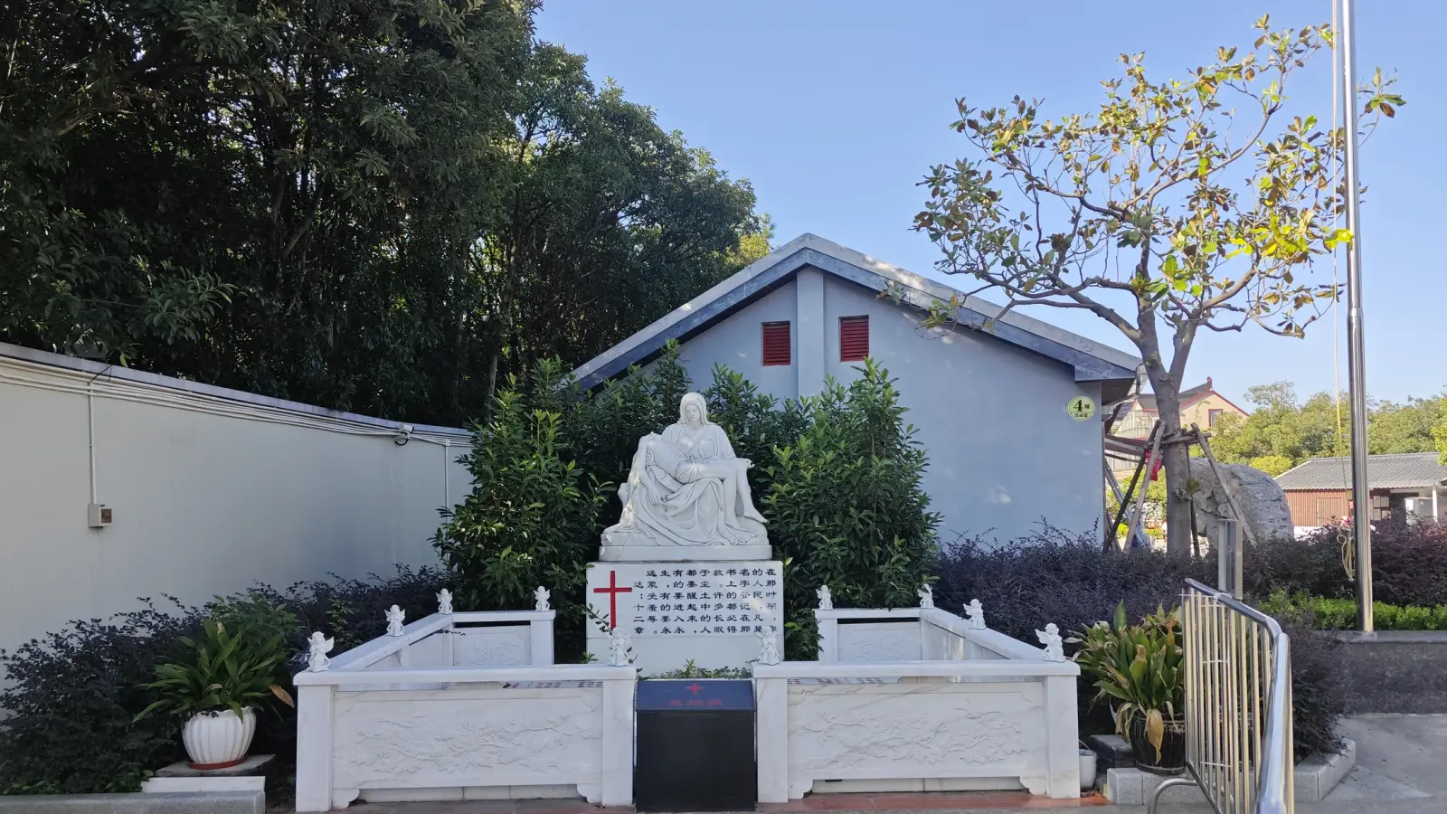 A white sculpture of Mary holding Christ inside a white marble railing, with a small building and bare tree behind.
