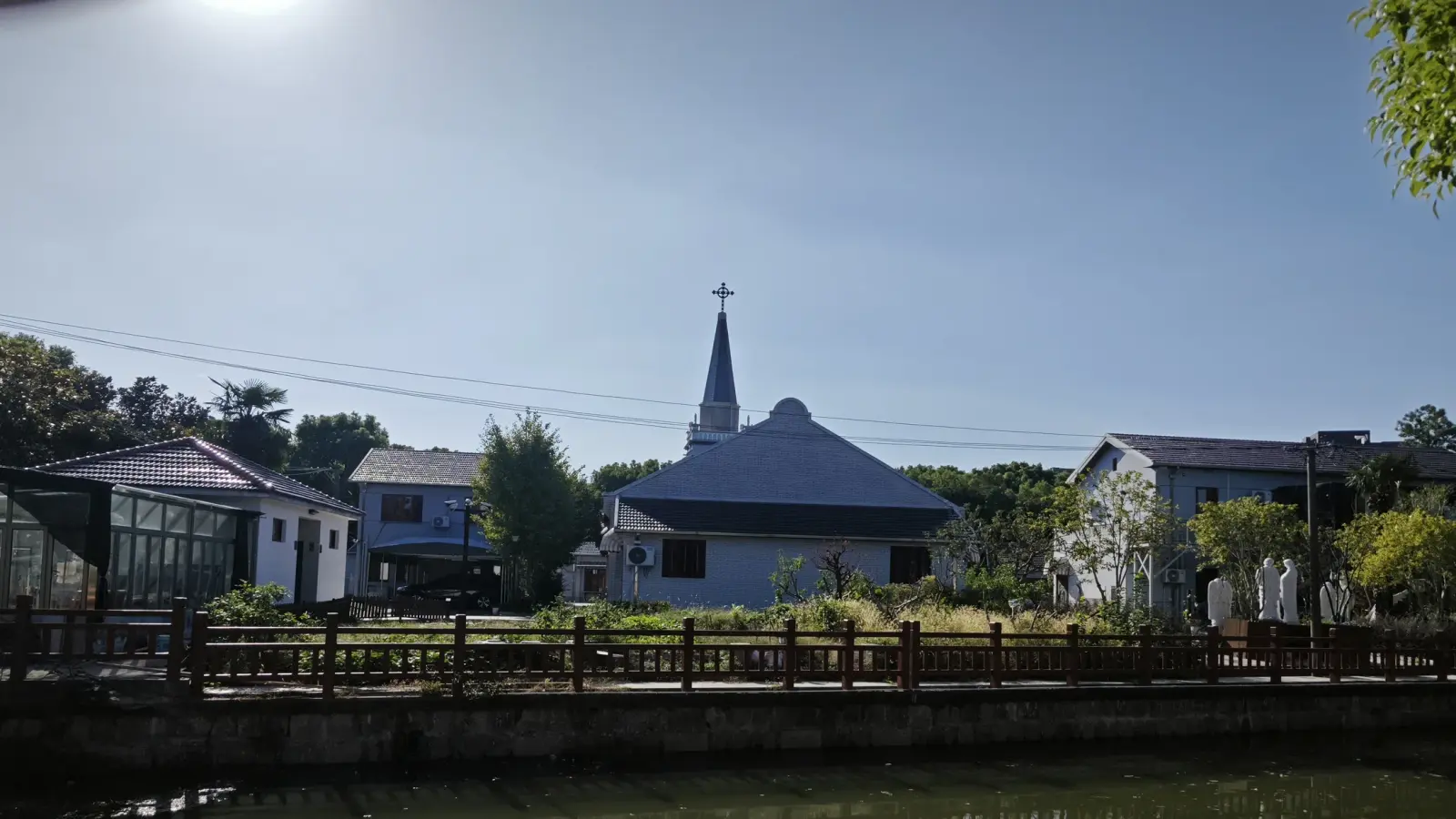 A dark grey roof and thin spire above low houses beyond a wooden railing and water.