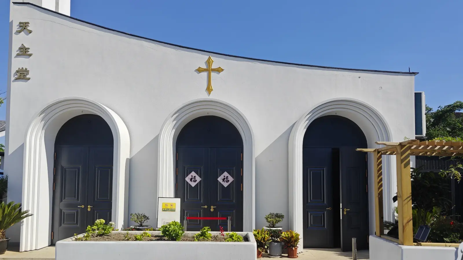 A white church front with three rounded-arch dark doors, a gold cross on the curved gable and Chinese characters down the left edge, in bright sunshine.