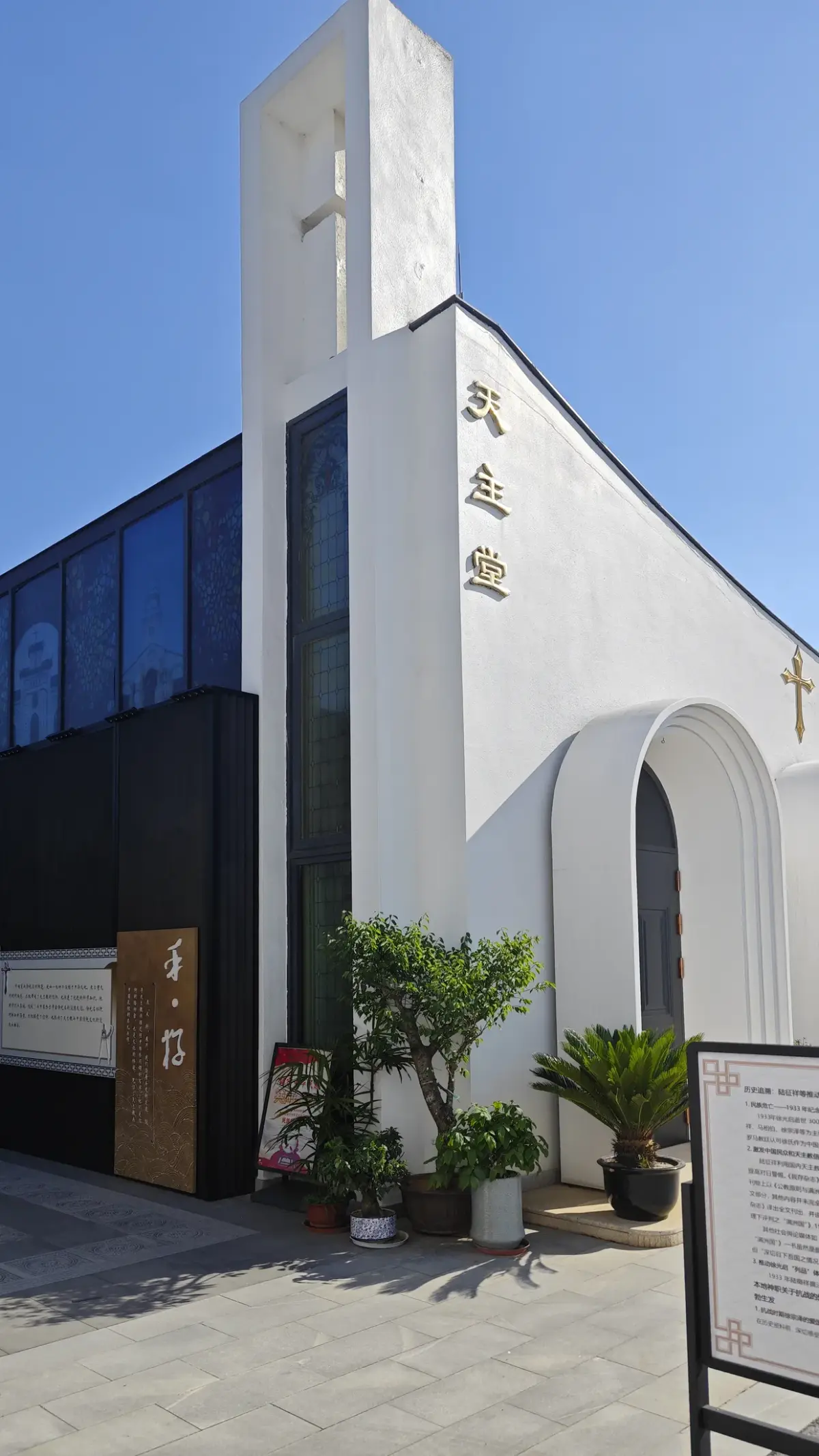 A white church wall with a tall narrow tower bearing a cross, a dark glazed side and potted plants at its foot.
