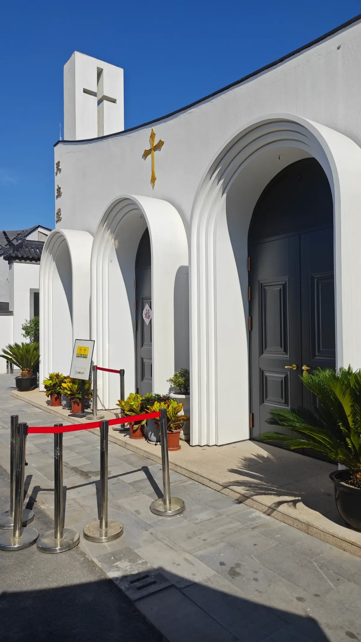 A white arched doorway with a gold cross above, a red rope on posts and potted plants.