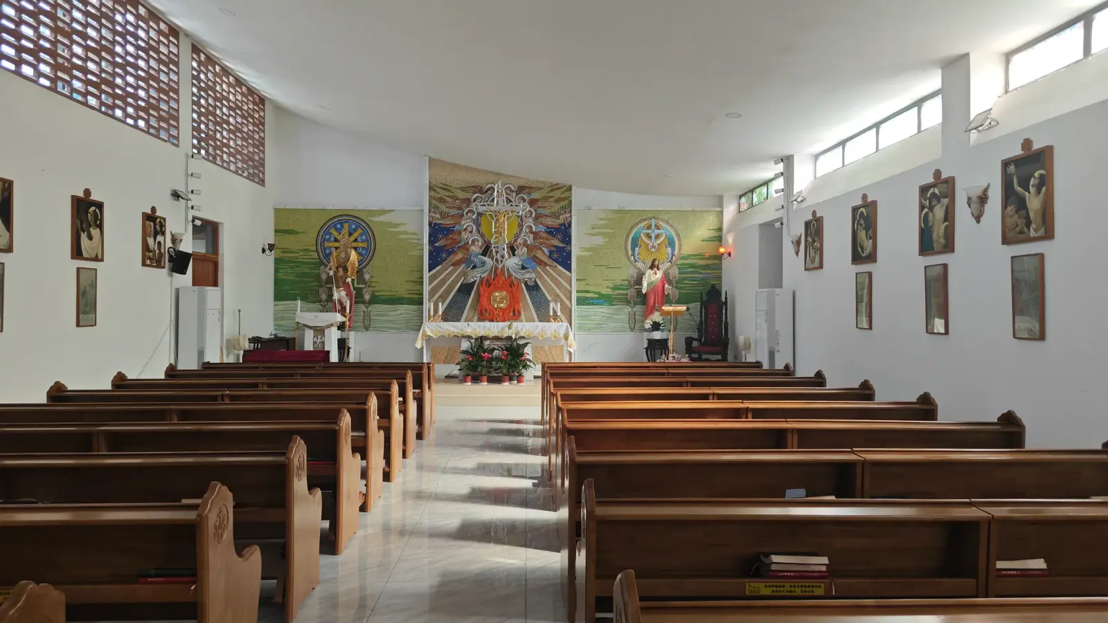 A bright white hall with long wooden pews, framed pictures on the walls and colourful panels at the far end.