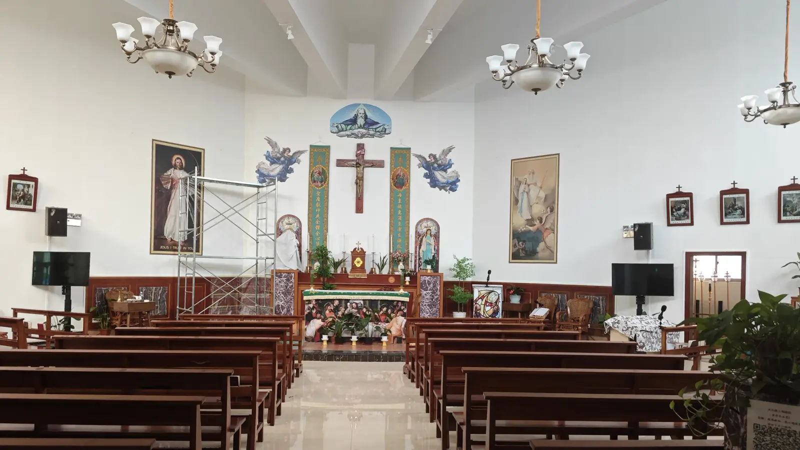 A bright church interior with a white wall, a central wooden cross, framed pictures, chandeliers and rows of dark wooden pews.
