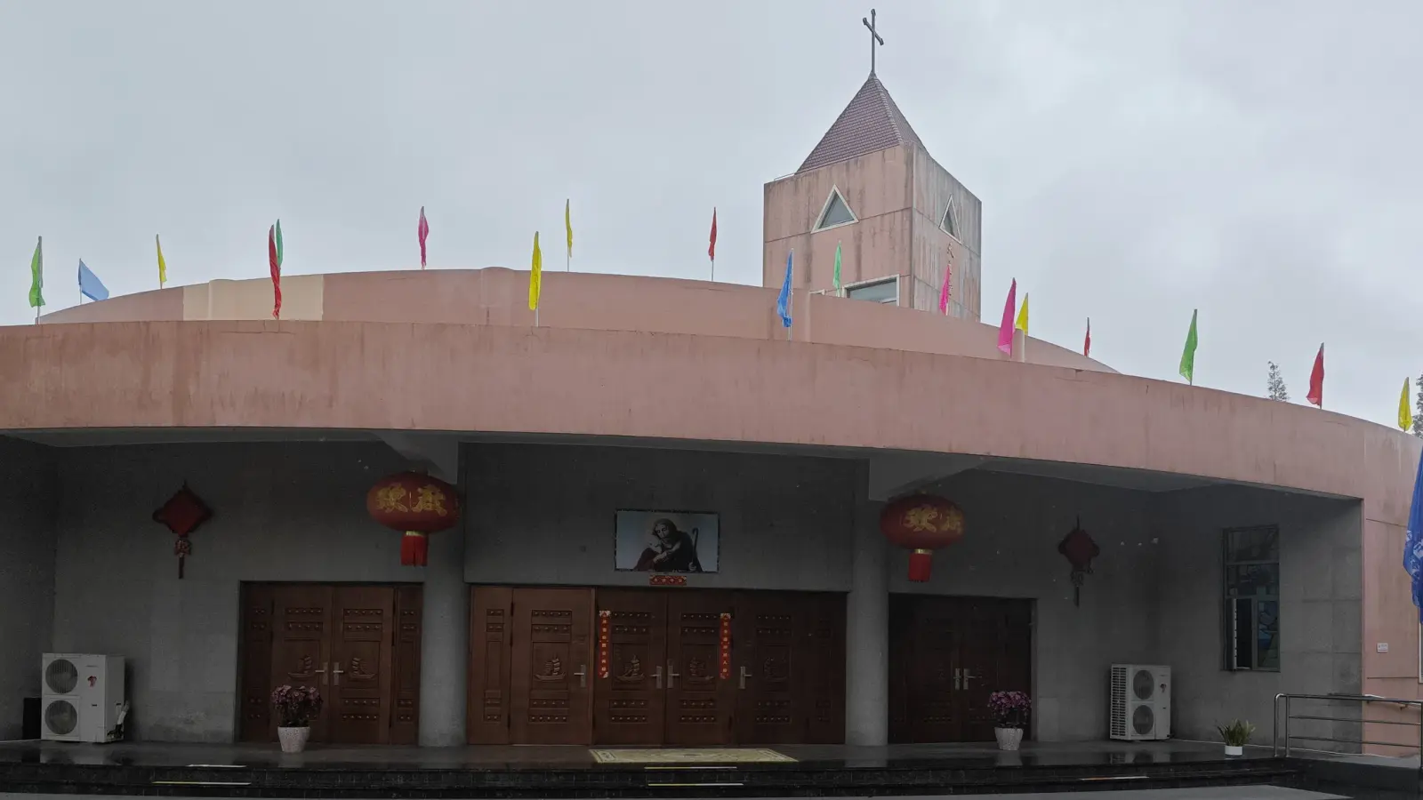 A pink curved-front church building with three brown wooden doors, red lanterns, a small tower with a cross on the roof and strings of coloured flags, under a grey sky.