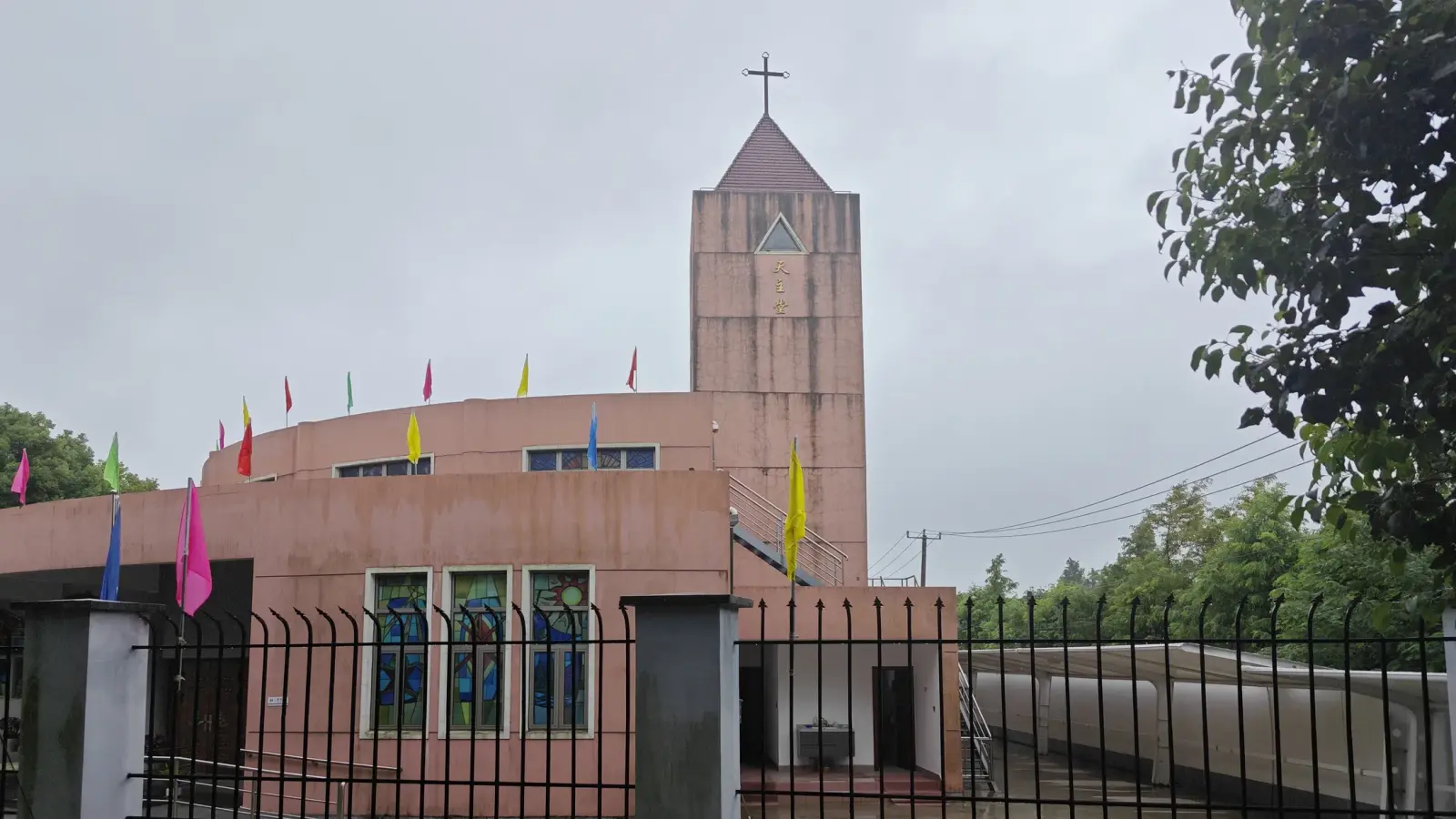 A pink brick tower with a cross, beside a curved pink building with three narrow stained glass windows behind a railing.