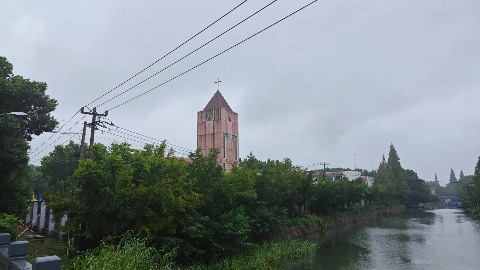 A pink-brown tower with a cross above trees beside a river under a grey sky.