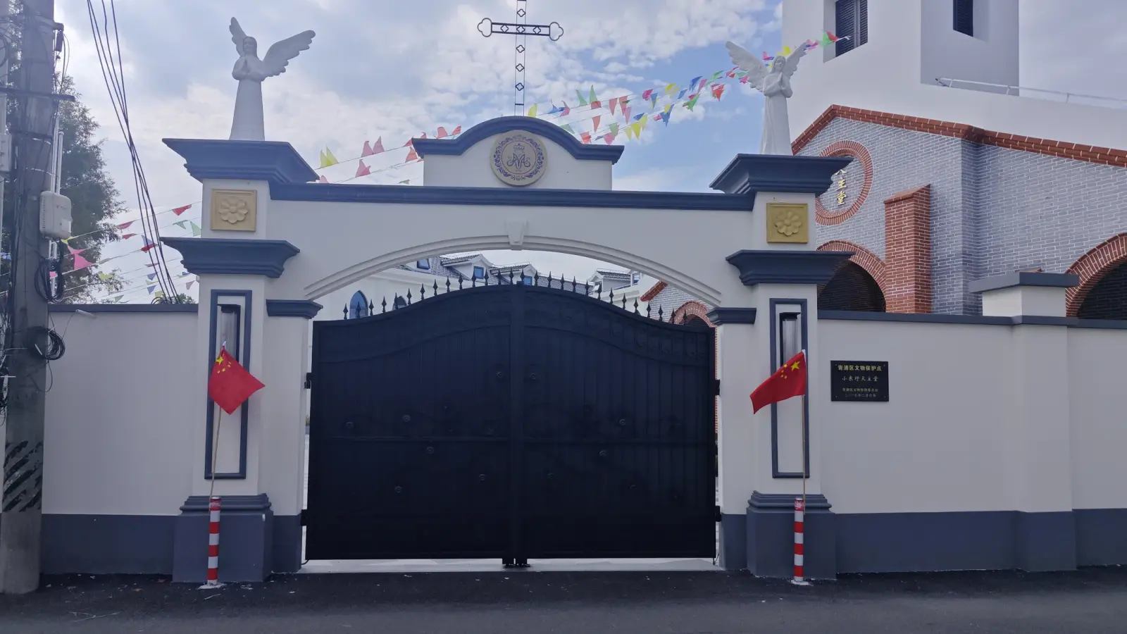 A wide black gate beneath an arched pediment with winged angel statues, flanked by red flags.