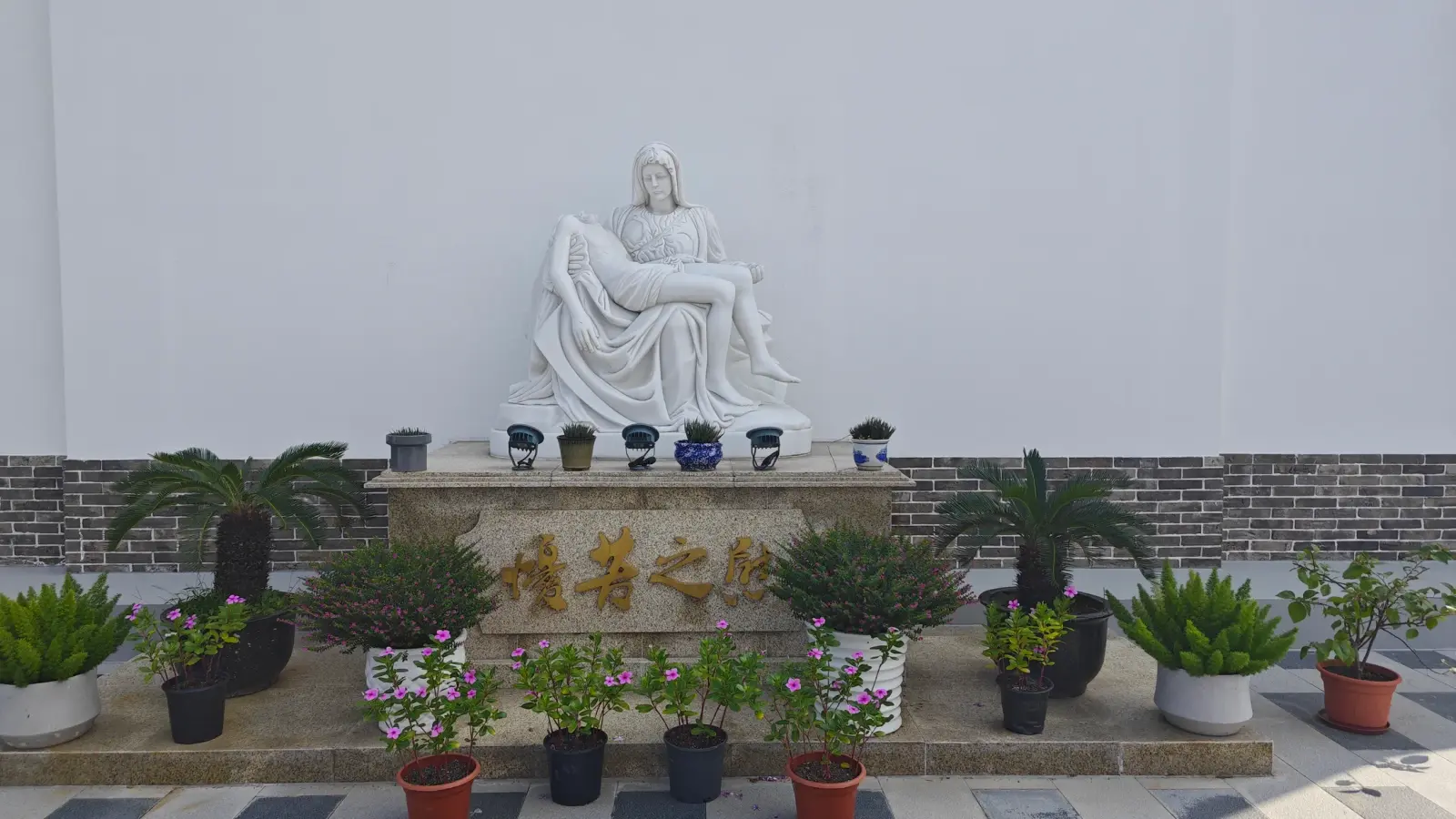 A white statue of Mary holding Christ on a stone plinth with Chinese characters, surrounded by potted plants.