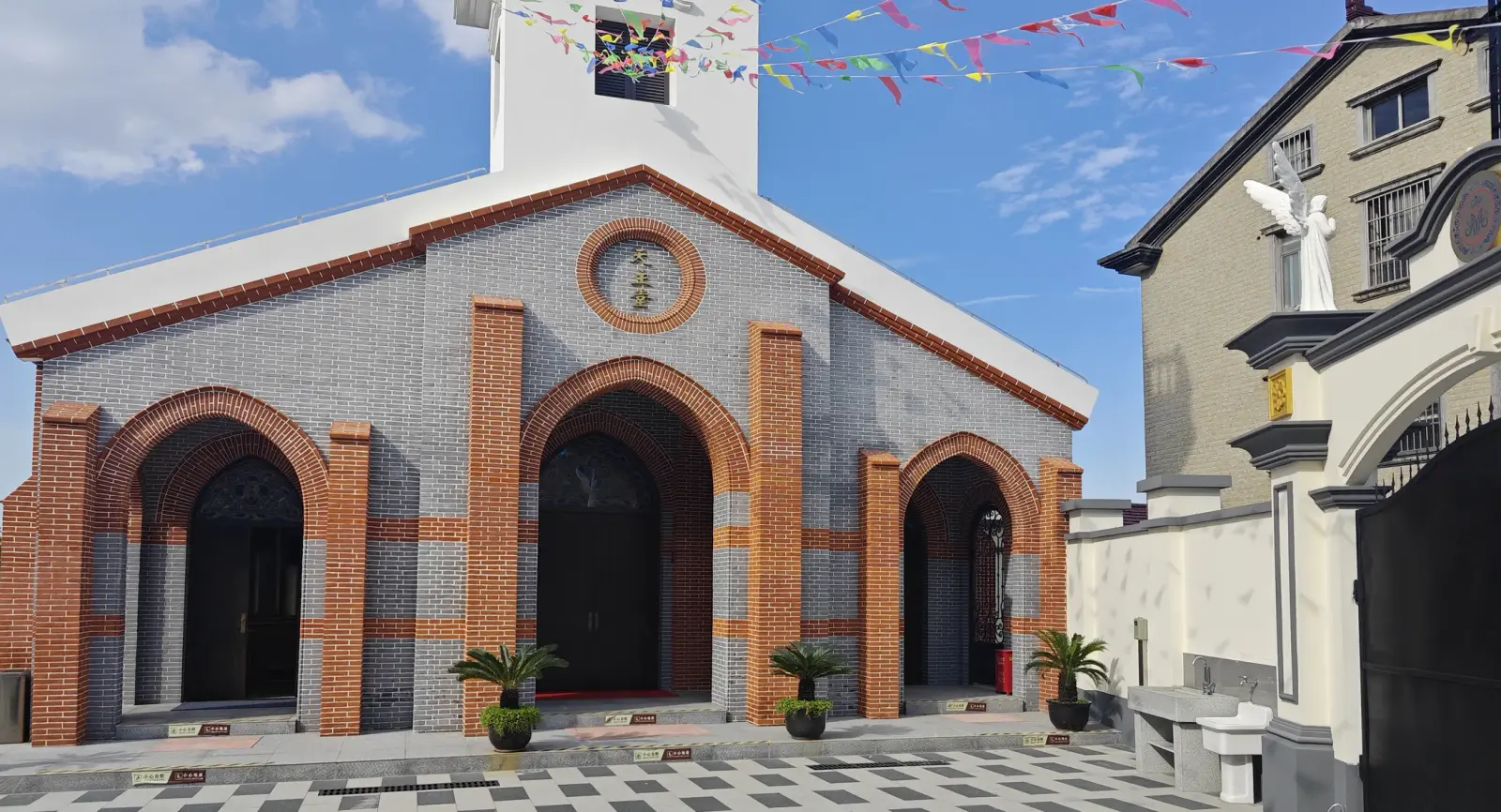 A church front in grey brick trimmed with orange-brick arches around two doorways and a round window, below a white bell tower, with coloured bunting overhead.