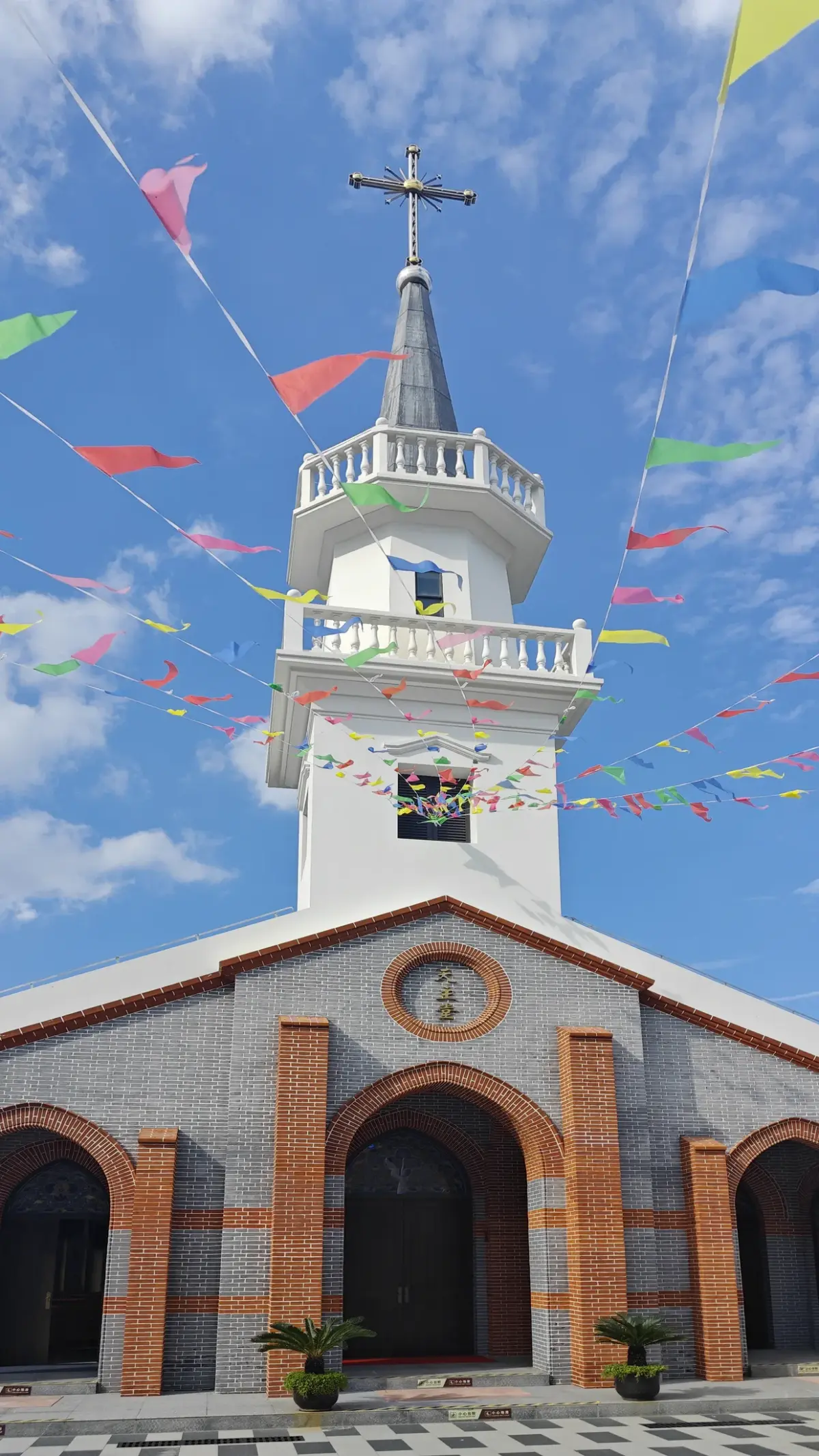 A white bell tower with a balcony and pointed spire above a brick-trimmed church front, under strings of coloured flags.