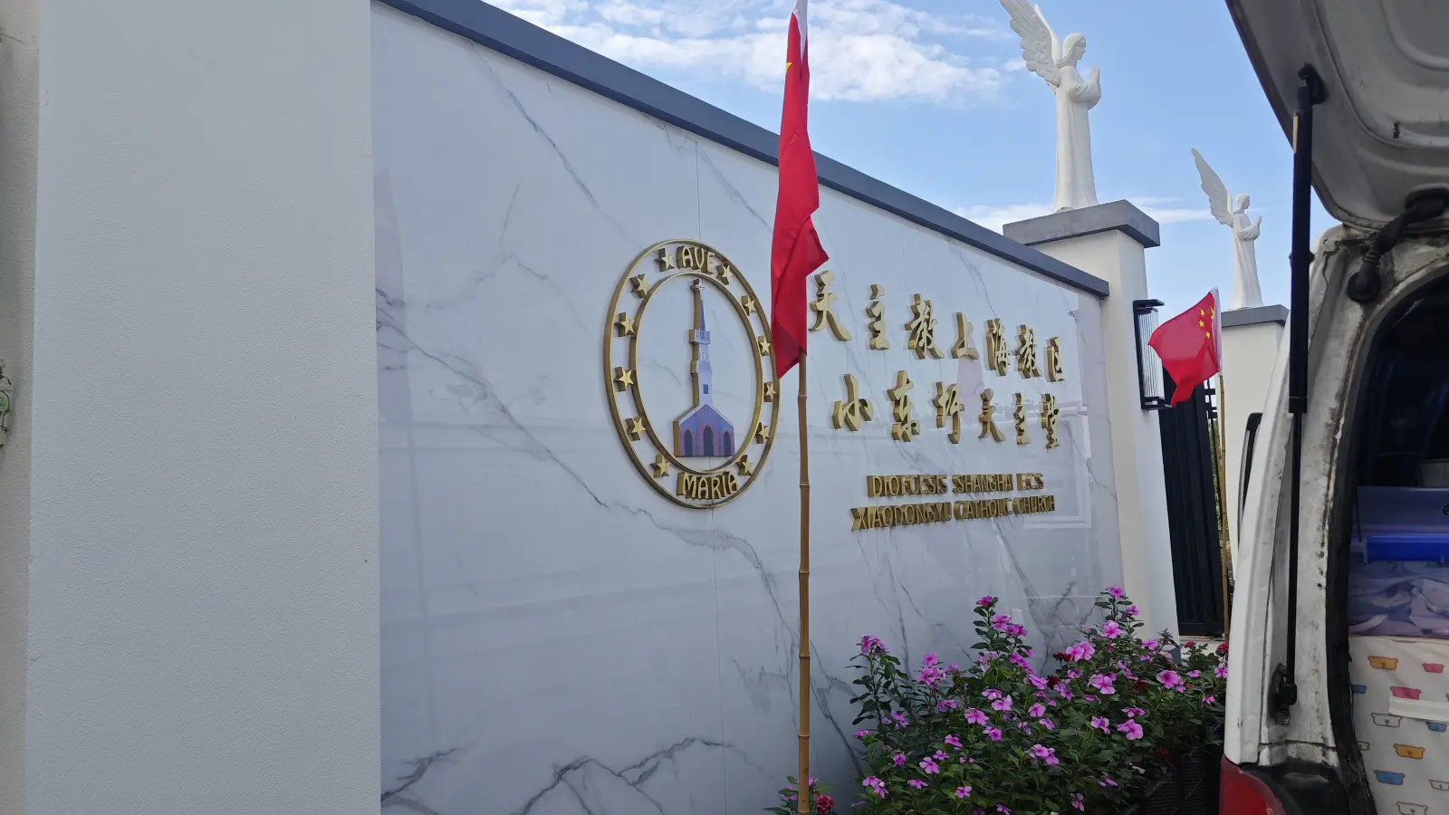 A white marble wall with a gold ring around a picture of a spired church, gold Chinese characters and English lettering, beside a red flag.