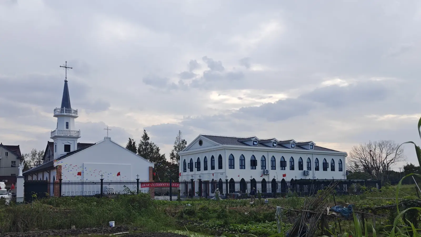 A white church with a tall spire next to a long white building with arched windows, beyond a field.