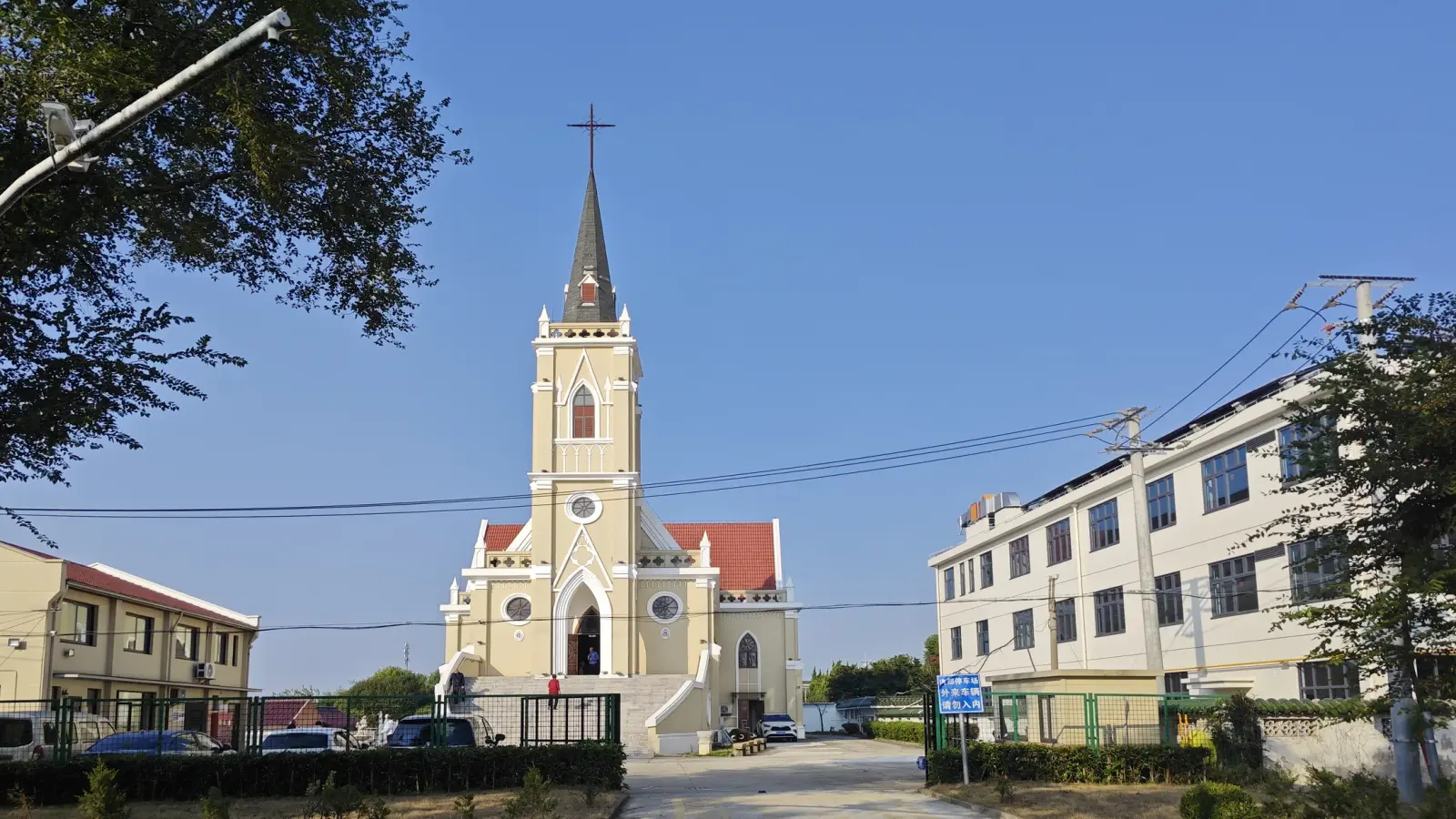 A cream church front with a high tower topped by a dark spire and cross, red roof tiles, and a plain building to its right, under a clear blue sky.
