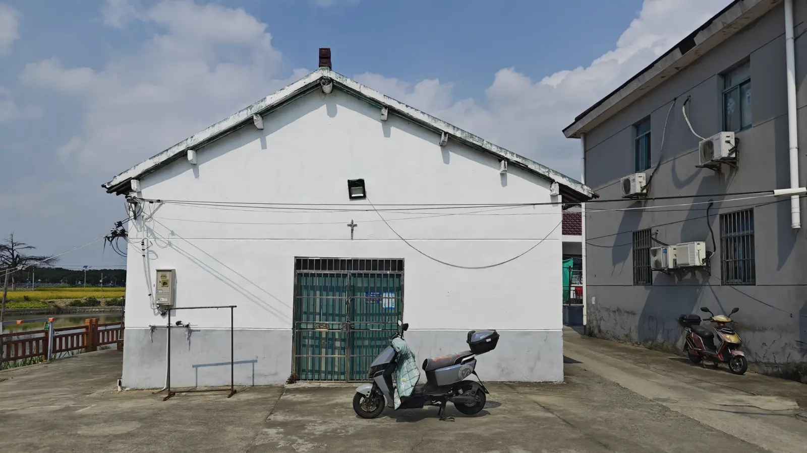 A single-storey white building with a gabled roof and a small cross above a green steel door, with scooters parked in front, under a blue sky.