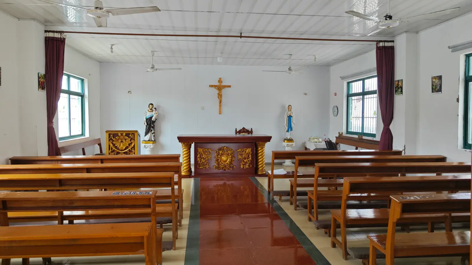 A plain white room with rows of wooden benches, a wooden crucifix, an altar table and two small statues, under a tiled ceiling.