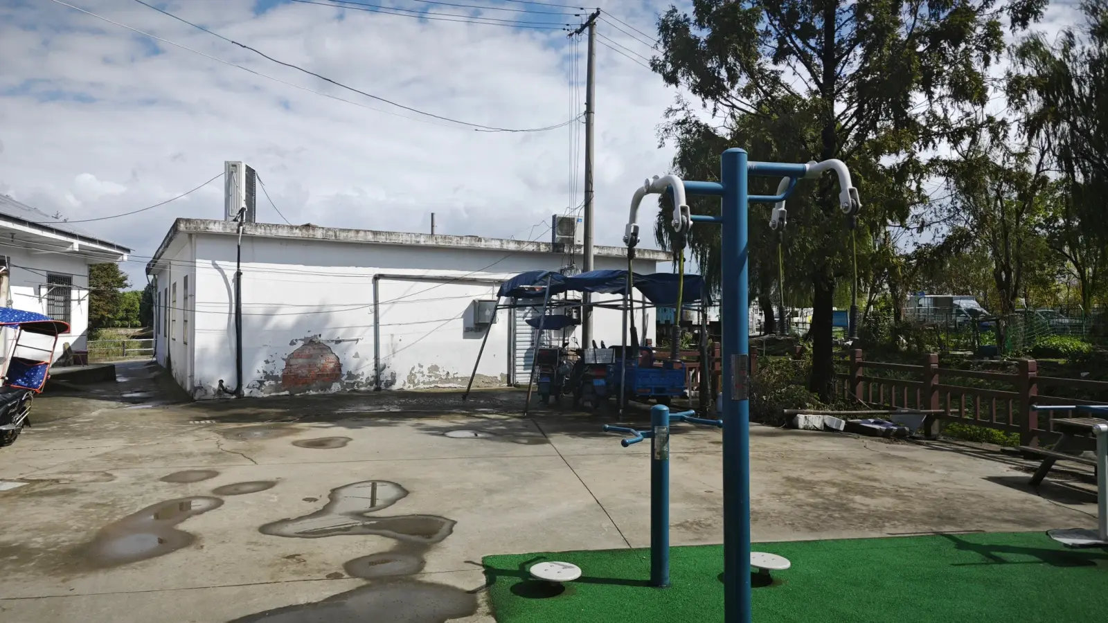 A low white building with a sloped roof behind a concrete yard, green mat and blue exercise equipment.