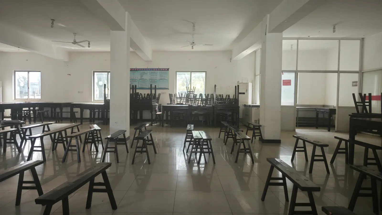 A wide white room with square pillars, wooden benches and tables arranged across a polished tile floor.