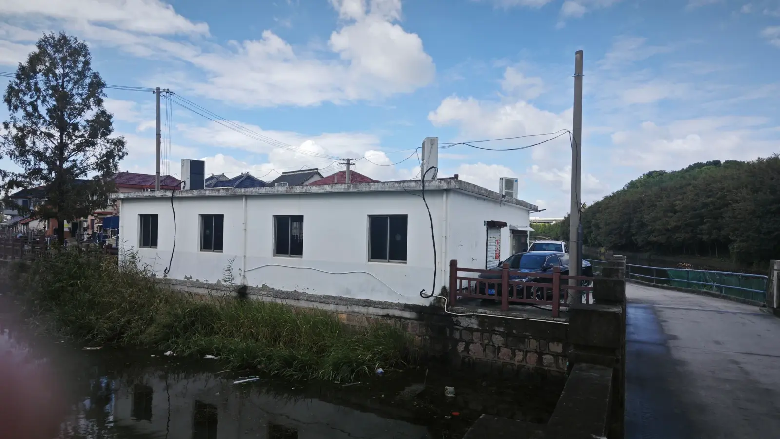 A single-storey white building with small windows beside a waterway and a paved lane, under a partly cloudy sky.