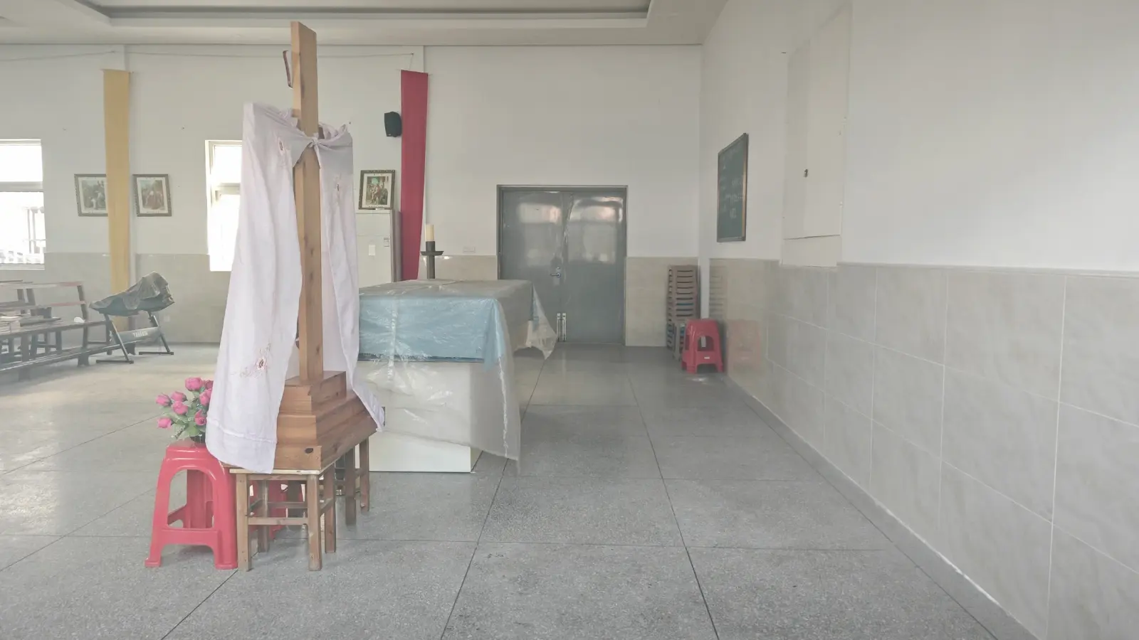 A plain white hall with a cloth-draped wooden cross, an altar table and red plastic stools.