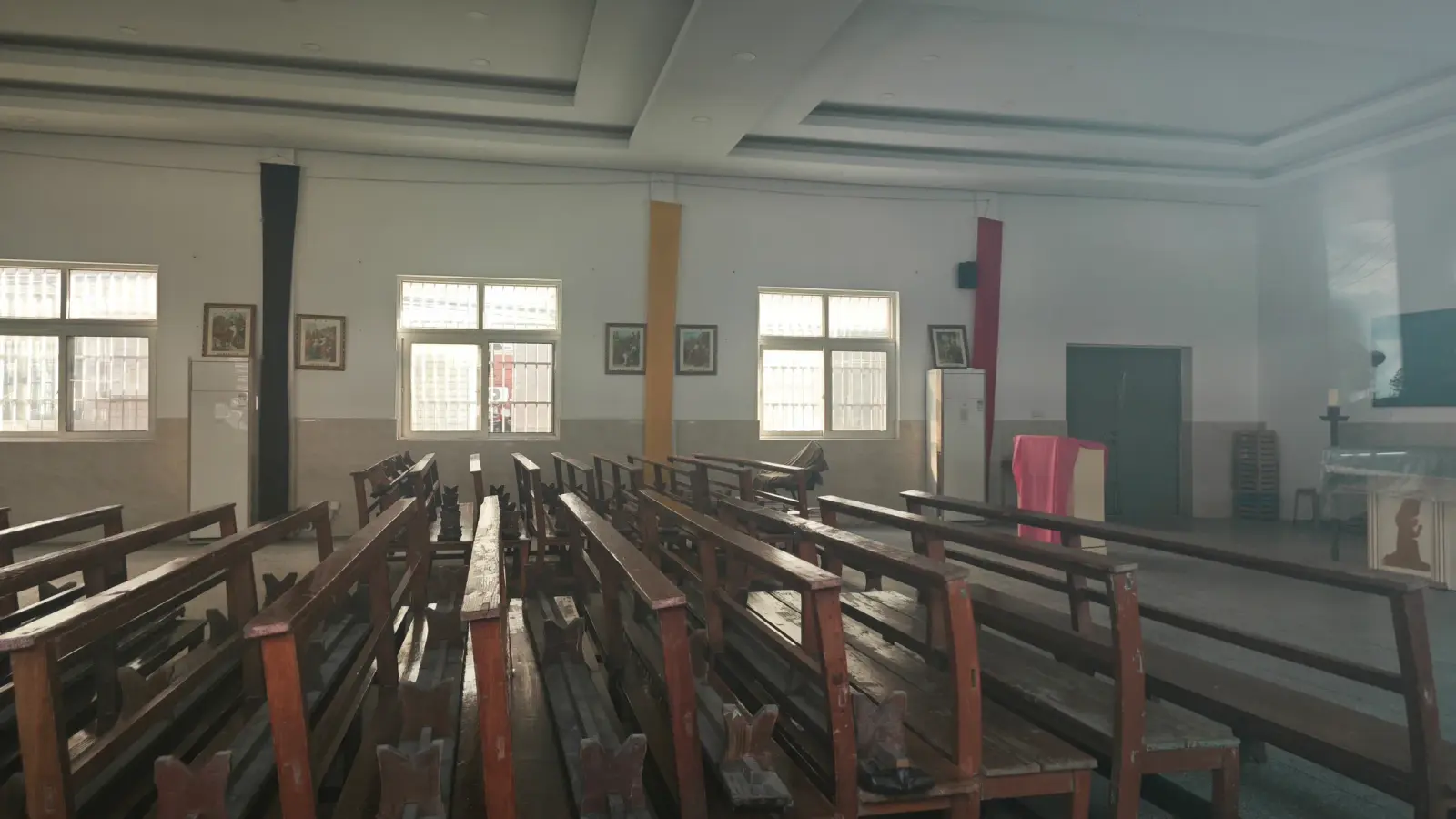 A room with long wooden benches, high windows and small framed pictures on the walls.