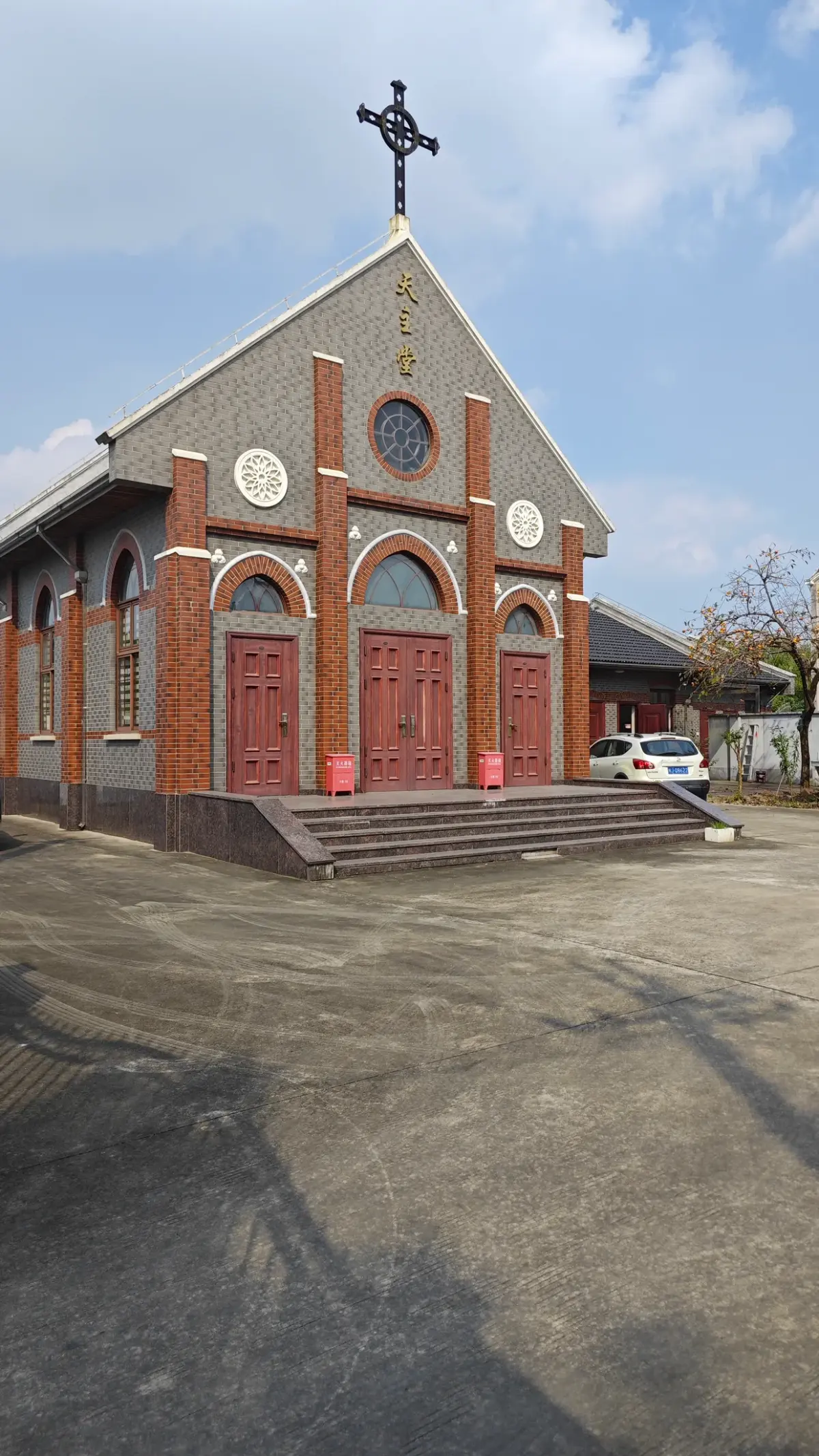 A gabled church front in grey brick with red-brick pilasters, a round window, a cross on top and red doors above low steps.