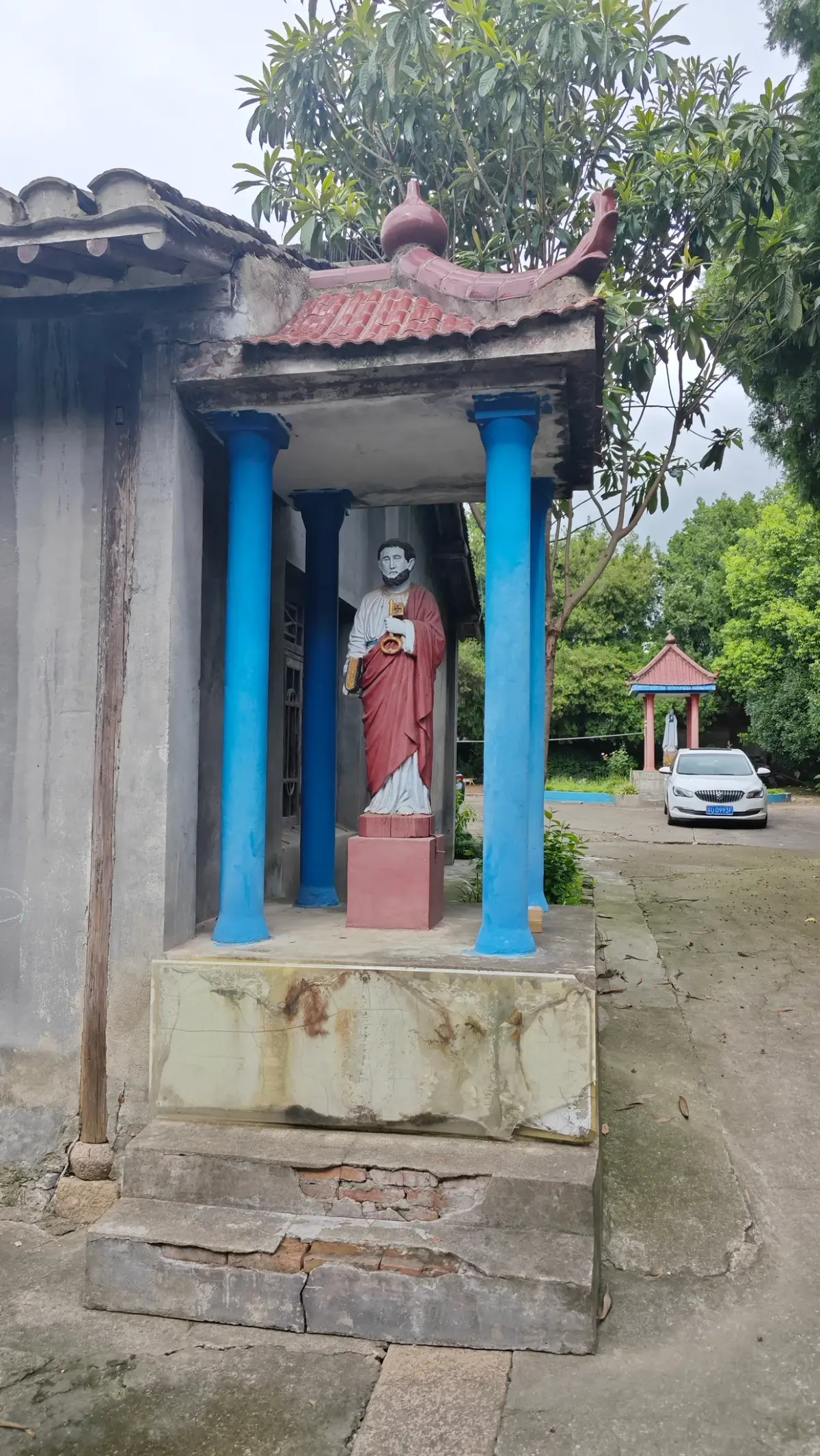 A statue of a figure in red and white under a small tiled roof on two blue columns, on a stone base.