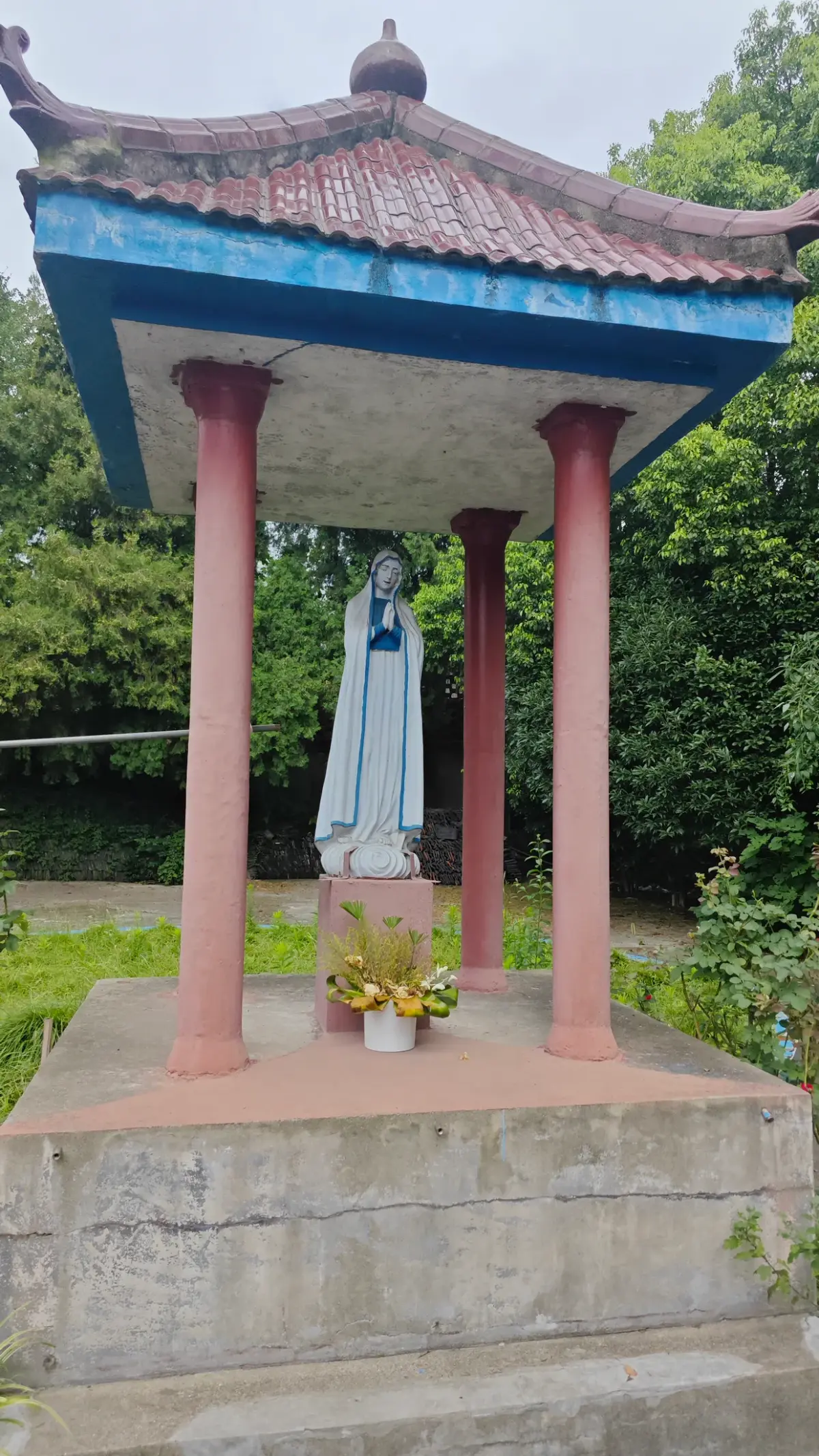 A statue under a blue-edged tiled pavilion roof on red columns, with trees behind.