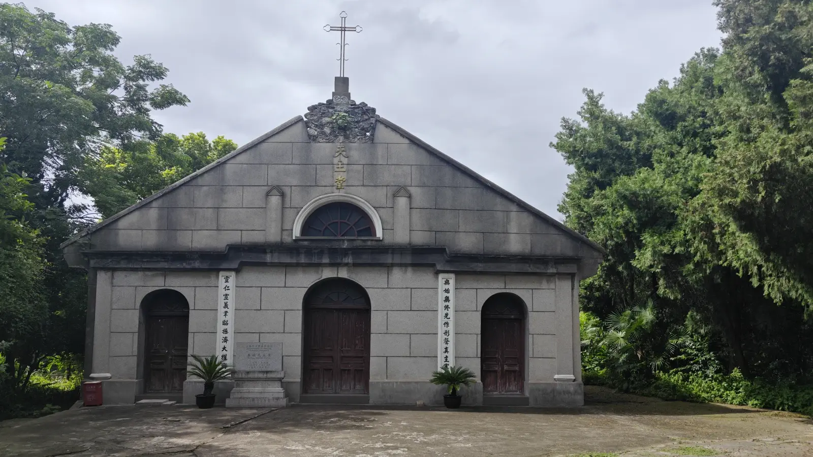 A grey stone church with a triangular gable, a round window, a cross on top and three arched wooden doors, framed by trees.