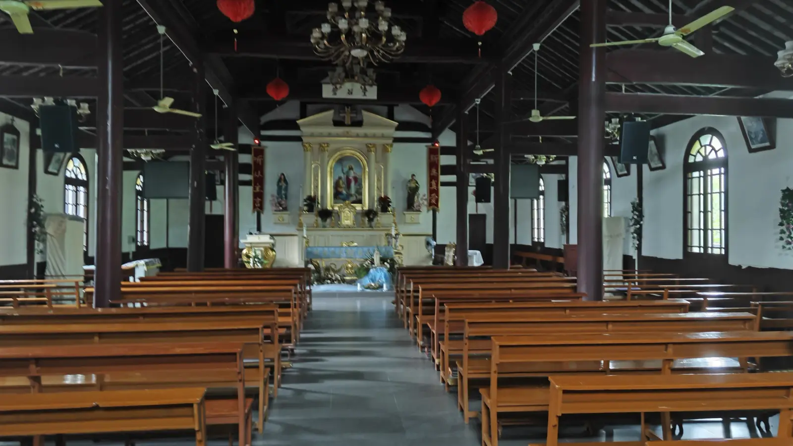 A church interior with exposed dark timber beams and columns, red hanging lanterns, long benches and a gilded altar at the end.