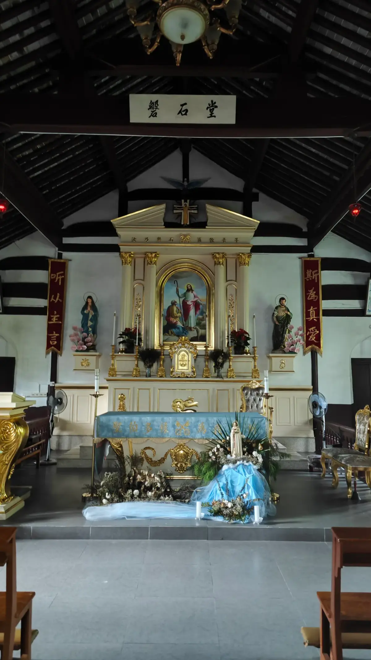 A gilded altar with a framed picture, flanked by red vertical banners, beneath dark roof timbers.