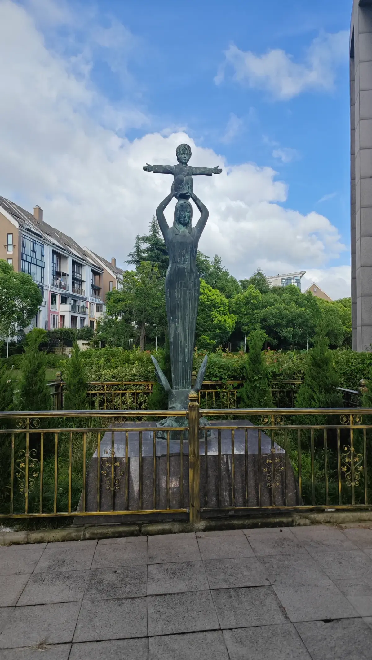 A dark statue of a standing figure lifting a smaller figure aloft, behind a gold-coloured fence.
