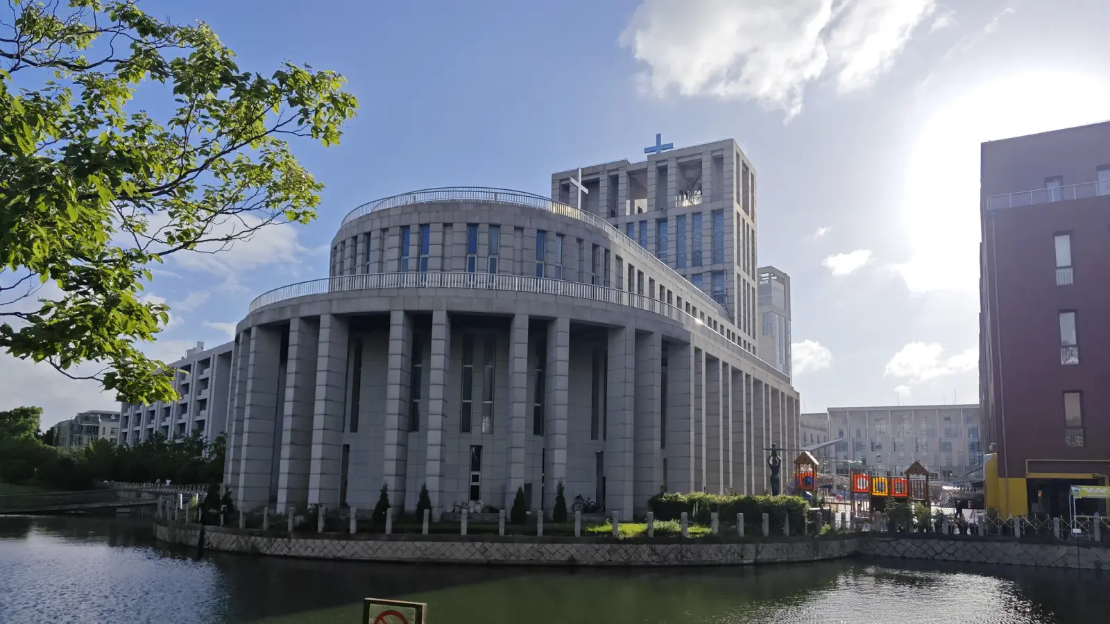 A modern stone church with a curved ring of tall columns and a tower behind, reflected in a lake in the foreground.