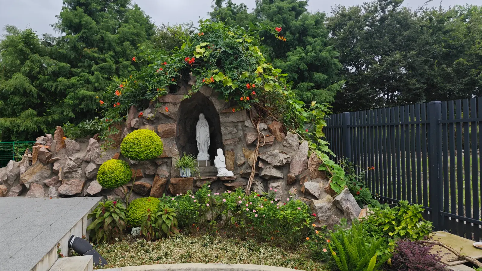 A rough stone grotto holding a white statue, framed by green shrubs and orange flowers.