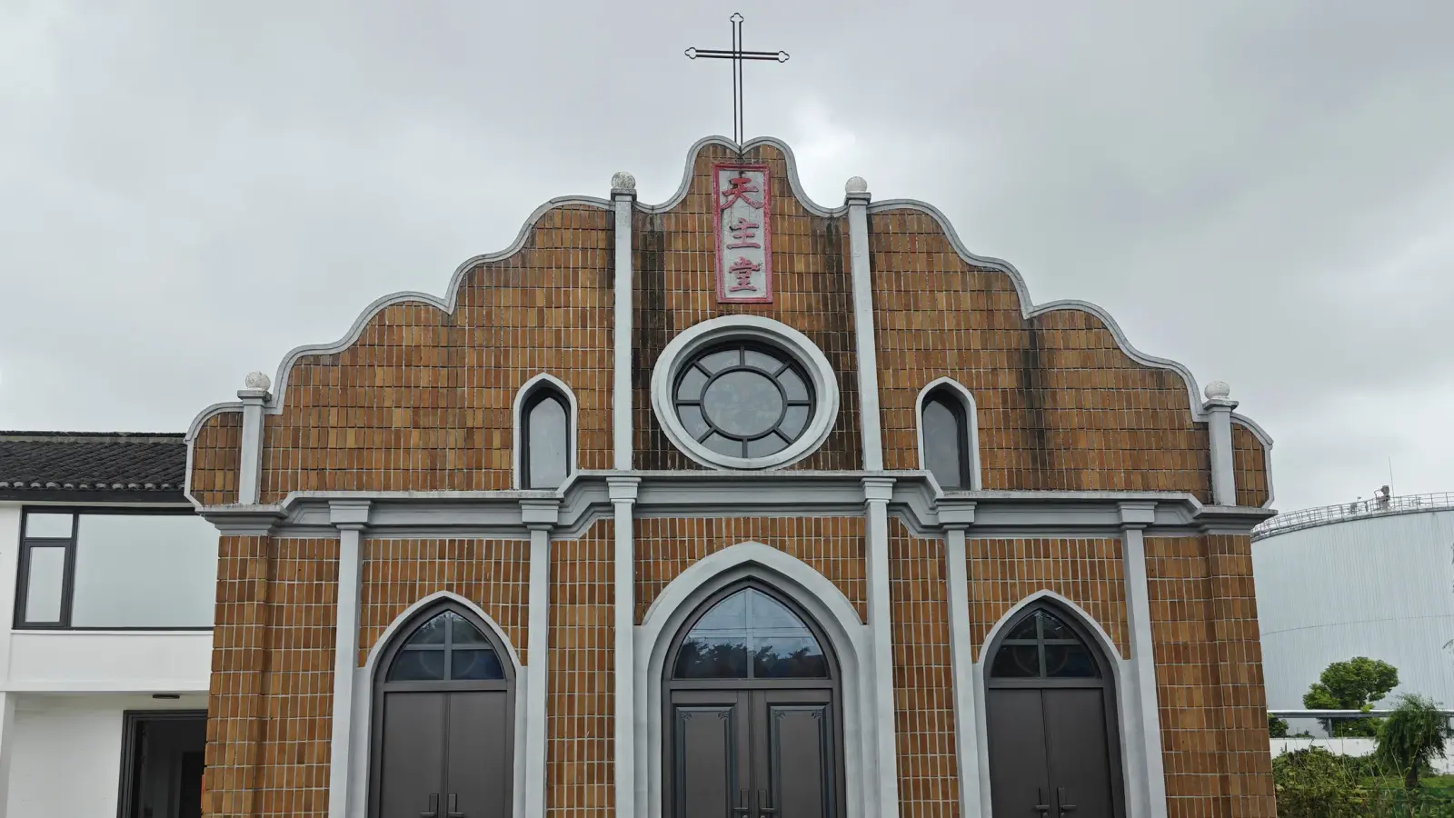 A church front clad in orange-brown scale tiles, with a stepped gable, a round window, a cross on top and three arched doors.