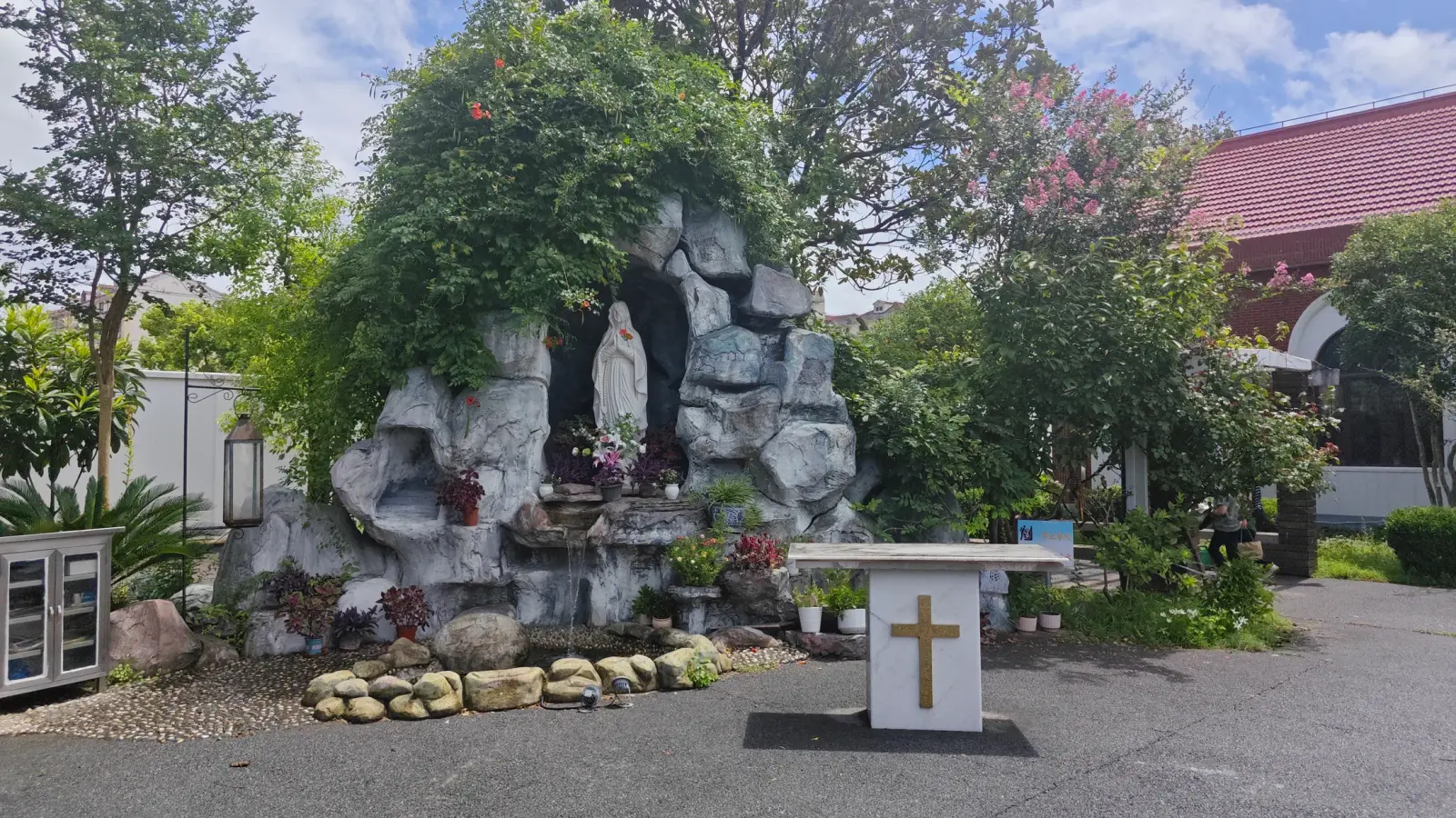 A grey rock grotto covered with climbing plants and a statue inside, with a white altar bearing a gold cross in front.