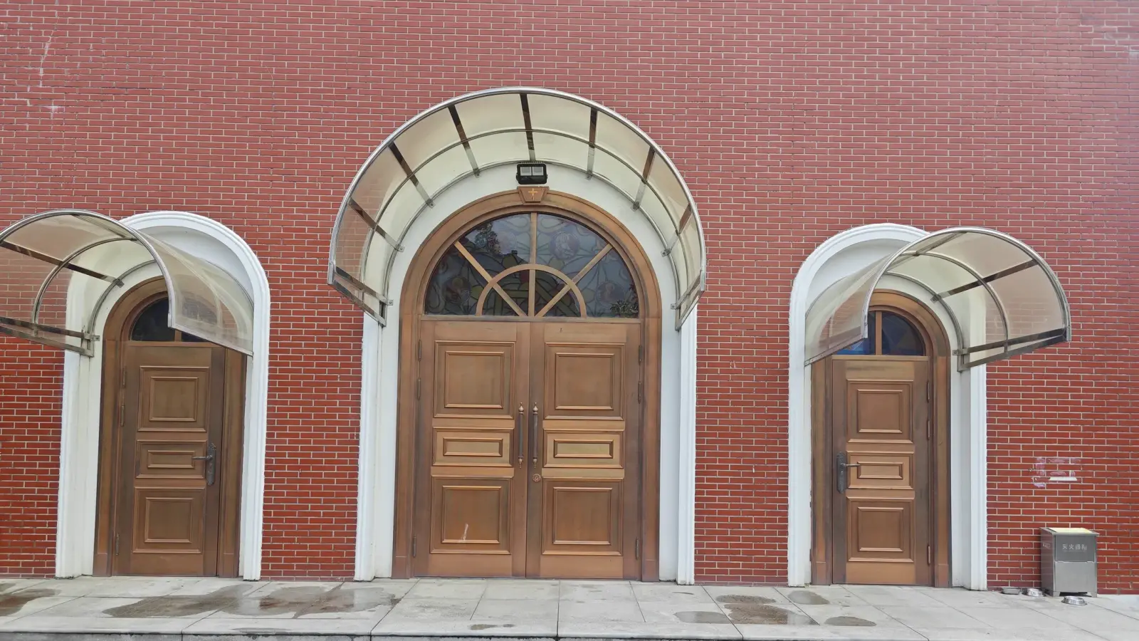 Three arched wooden doors set in a red brick wall, framed in white with glass canopies above.