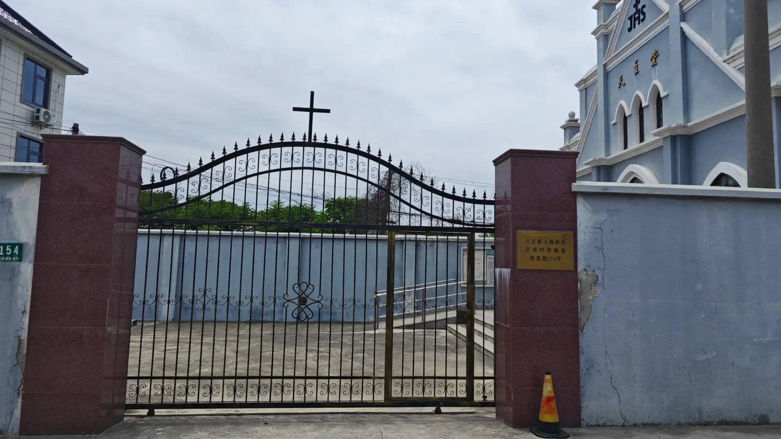 An iron gate with a cross between two maroon pillars and a pale blue wall, with a blue church gable behind.