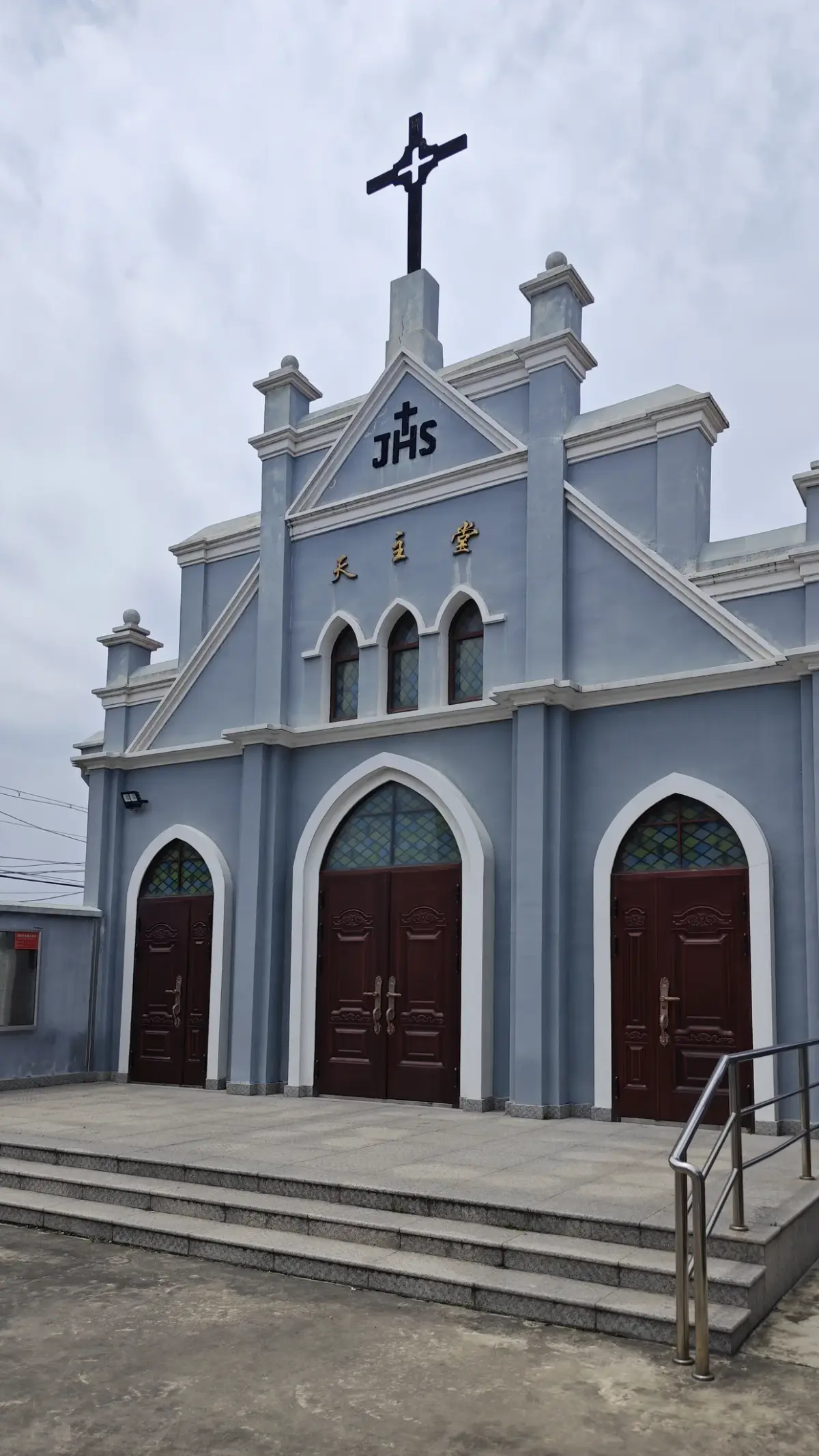 A pale blue church front with a stepped gable, a cross at the top, small arched windows and three white-framed arched red doors above concrete steps.