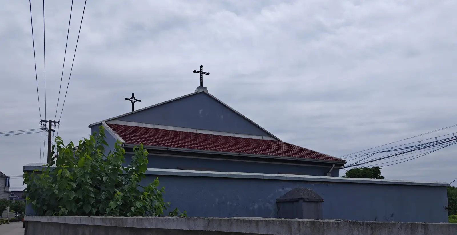 A blue church wall with a red tiled roof and two crosses on the ridge, behind a low grey wall.
